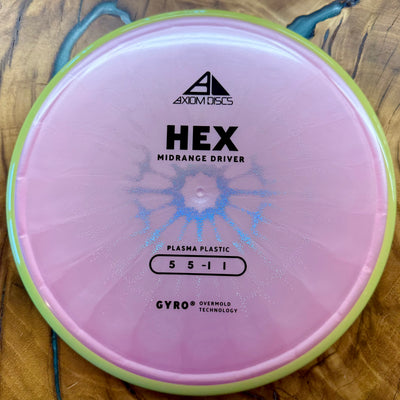 A pink Axiom Discs Hex midrange disc golf disc with a light green outer rim, photographed from directly above on a wooden surface.