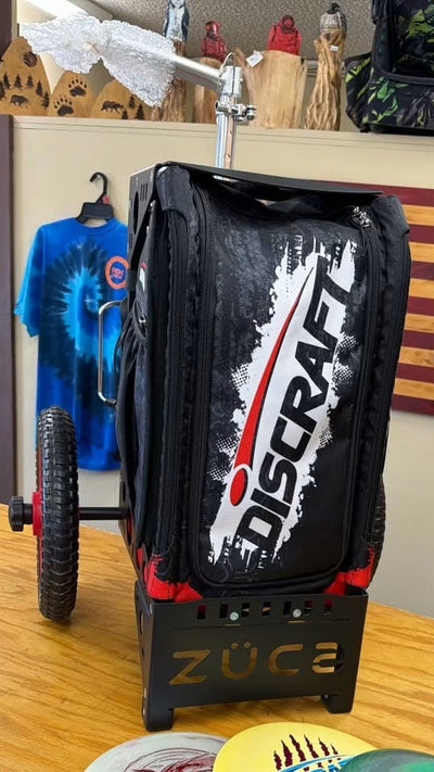 A black ZUCA Discraft disc golf bag with a wheeled cart base, standing on a wooden table, with three disc golf discs in front of it.