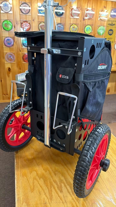 A black Discraft ZUCA disc golf cart with red-spoked wheels, shown on a wooden table in front of a wooden wall decorated with disc golf discs.