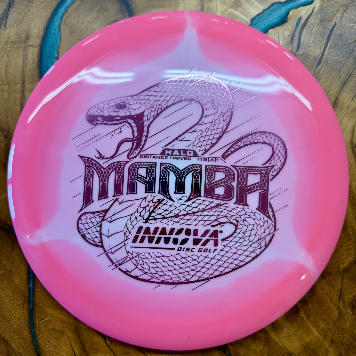 A pink Innova Mamba disc golf disc with a glossy finish, featuring a black snake graphic and the model name 'MAMBA' on a wooden surface.