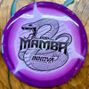 A purple and white swirled disc golf disc with a black printed snake graphic and the text 'Mamba' and 'Innova Disc Golf' on a wooden surface.