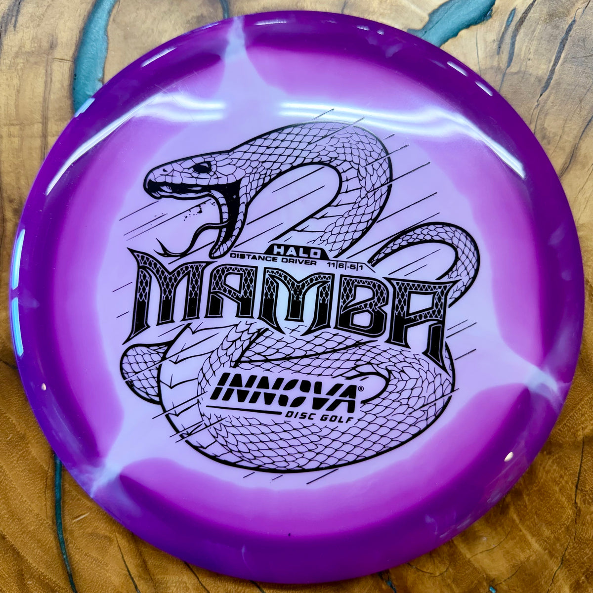 A purple and white swirled disc golf disc with a black printed snake graphic and the text 'Mamba' and 'Innova Disc Golf' on a wooden surface.