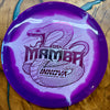 A purple and white swirl Innova Mamba disc golf driver disc, featuring a black snake graphic and text, resting on a wooden surface.