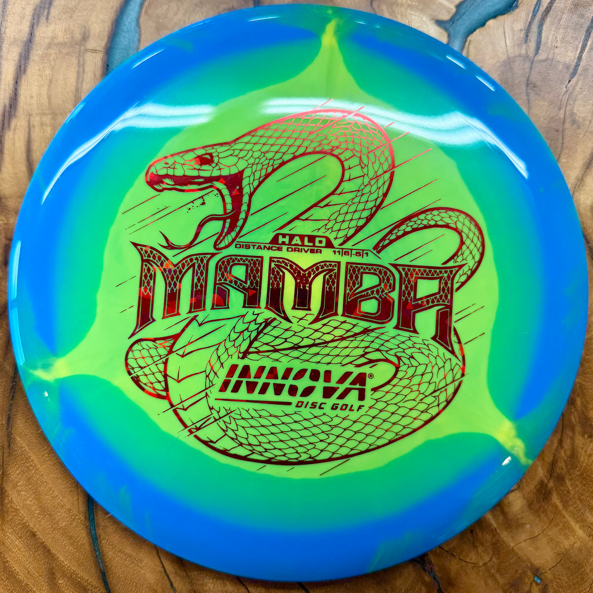 An Innova Mamba disc golf disc featuring a blue and neon green gradient design with a red snake graphic and the word 'MAMBA' in the center, shown on a wooden surface.