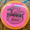 A pink and orange Innova Mamba disc golf disc with a black snake graphic and text, resting on a wooden surface.