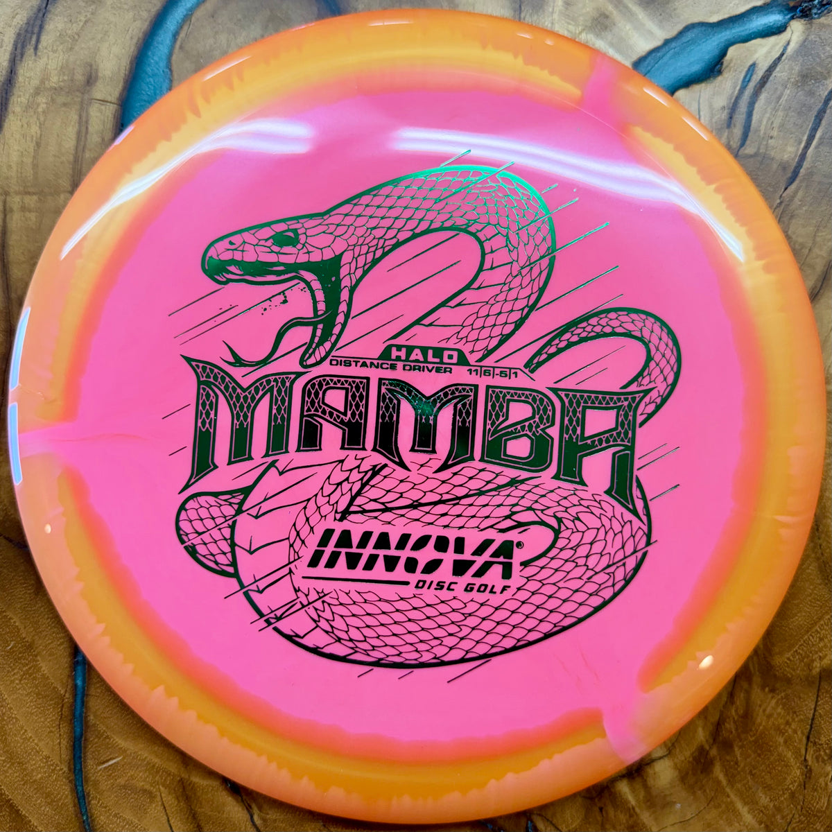 A pink and orange Innova Mamba disc golf disc with a black snake graphic and text, resting on a wooden surface.